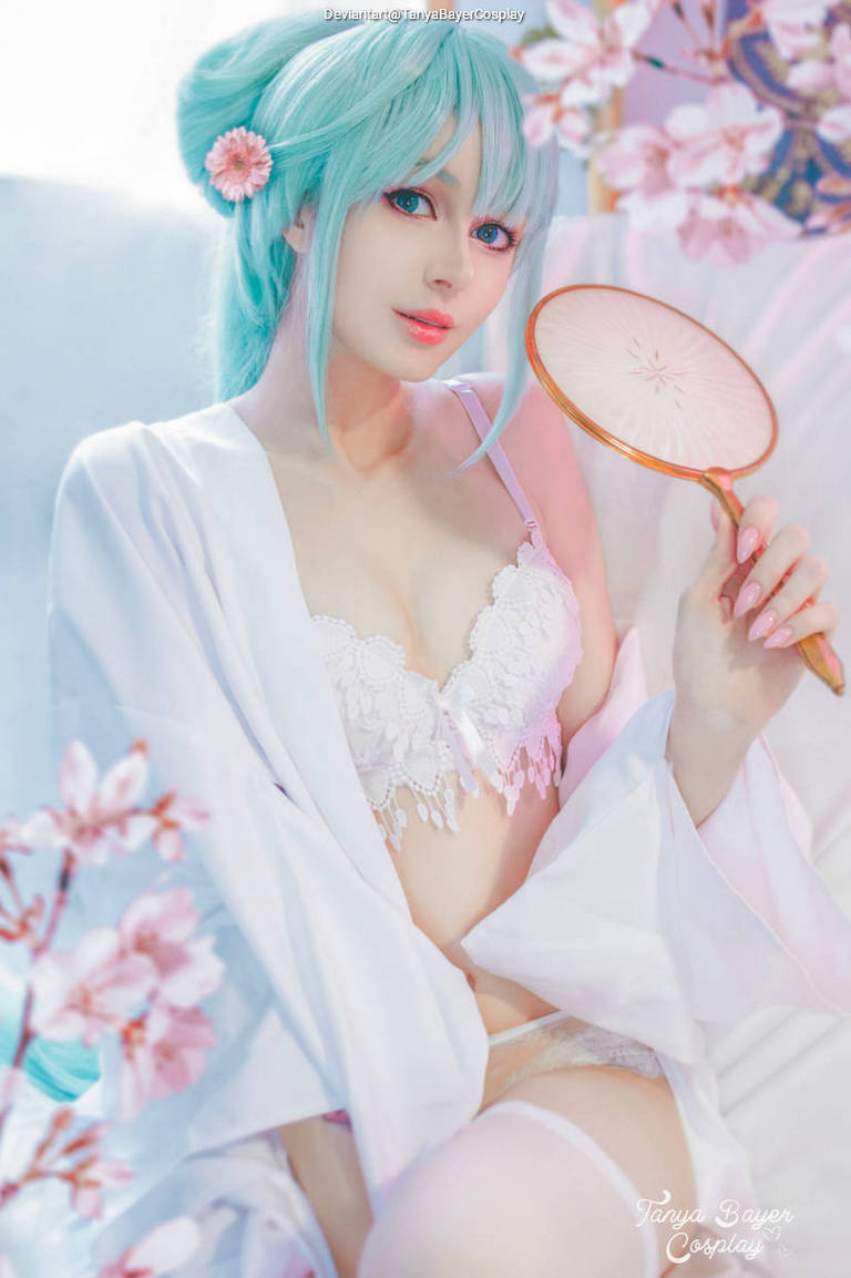 A female cosplayer with long turquoise hair and a floral accessory holds a traditional fan while wearing a white robe. The background features pink cherry blossom branches.