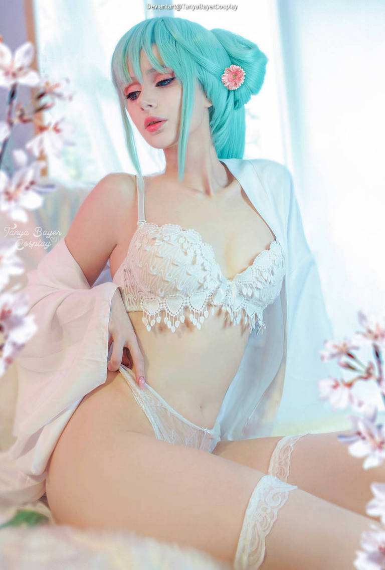 A woman with teal hair styled in a bun and bangs, wearing a white lace bralette and matching lace panties, is posing elegantly among white flowers and soft drapery.