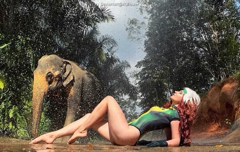 A woman in a green and yellow swimsuit lies on the ground in a lush, tropical setting while an elephant stands nearby, surrounded by splashing water and foliage.