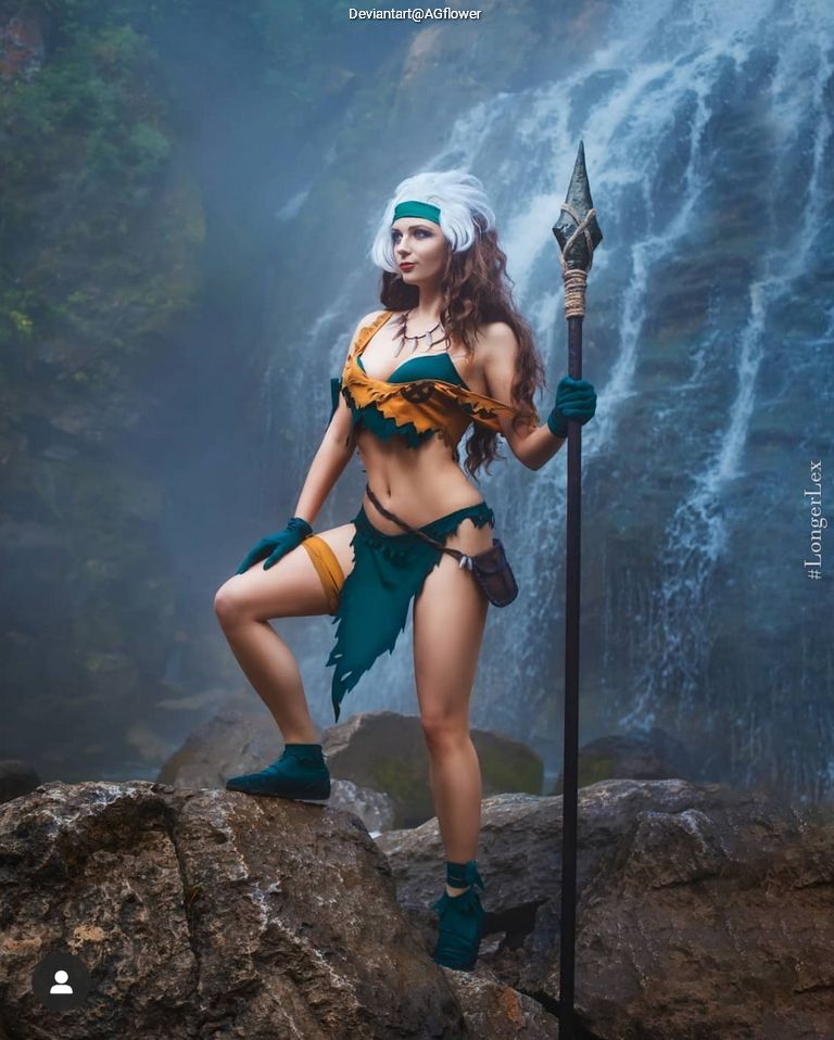 A warrior woman in a green and brown costume poses confidently on rocks near a waterfall, holding a spear.