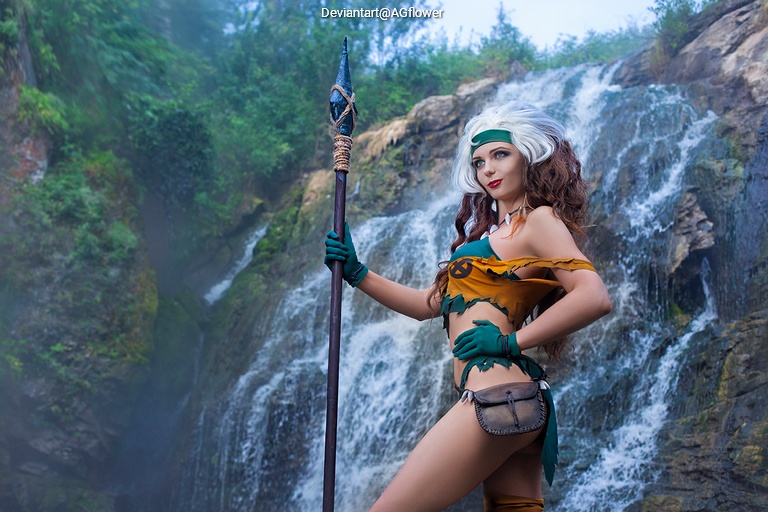 A female cosplayer poses confidently in front of a waterfall, wearing a colorful costume with green and beige accents and holding a spear.