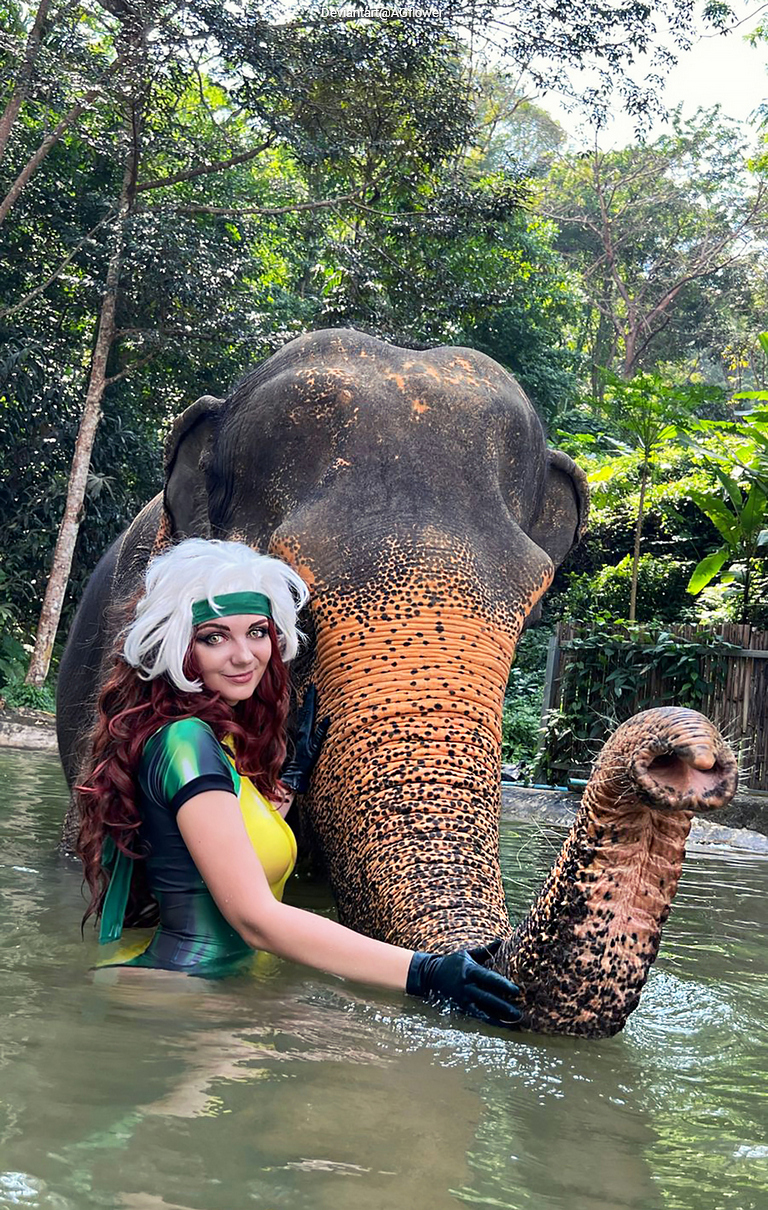 Person in a colorful outfit interacting with an elephant in a muddy water setting, surrounded by greenery.